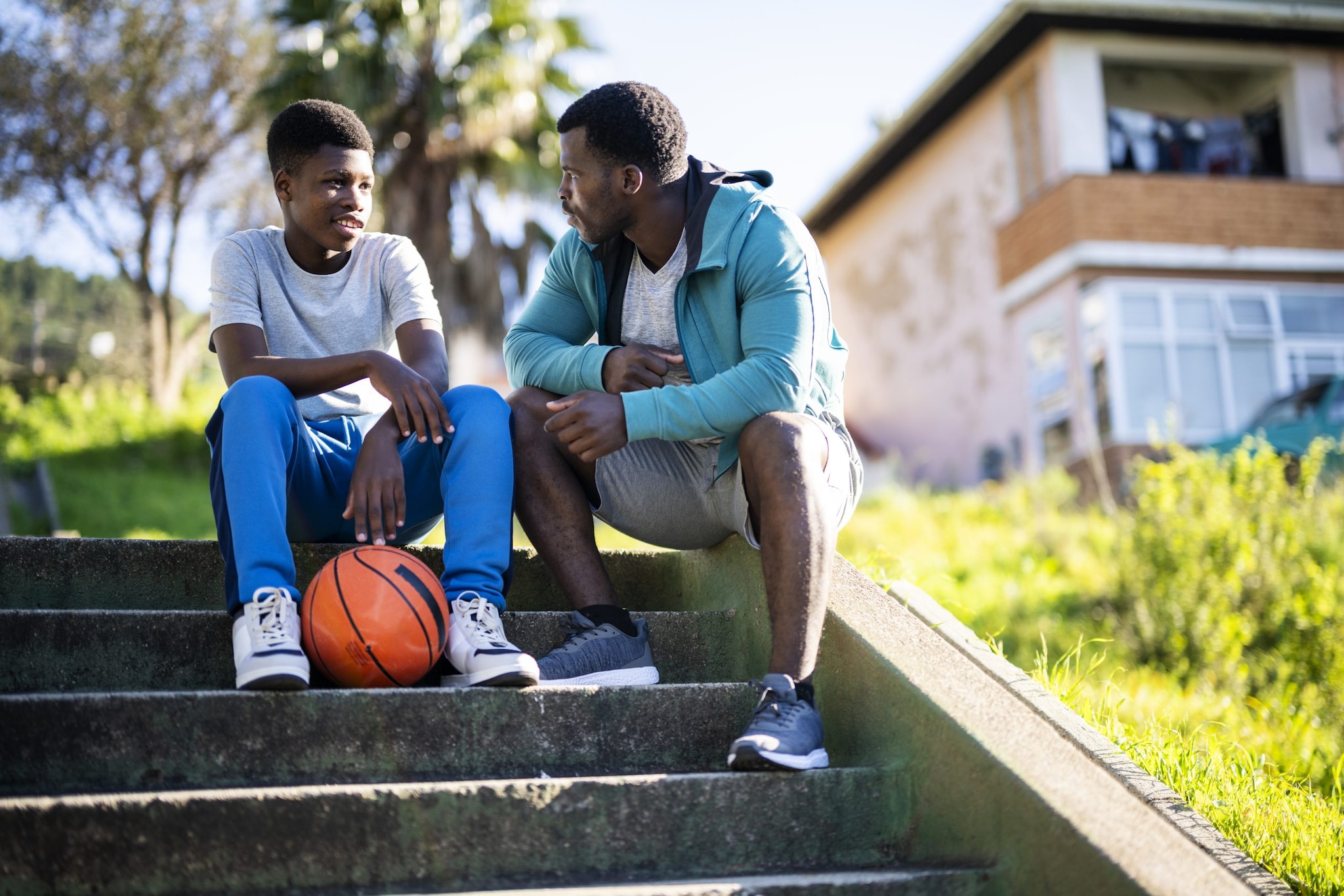A man and a child are sitting and talking on steps outside a housing area. A basketball lies at the feet of the young boy.
